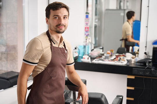 Amicable young man in apron working at hair salon