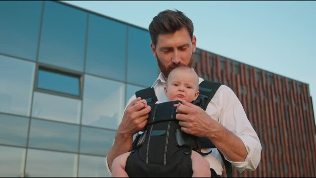 Cute portrait of funny newborn baby in a slings carrier. Young father holding a child kiss and hug on street. Ergo backpack. Parents. Slow motion