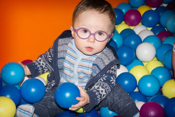 Fototapeta na wymiar A little boy in blue sweater with glasses plays with multi-colored plastic balls in the playroom. And holds two in his hands.
