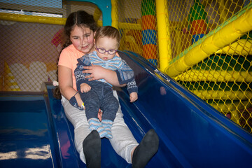 In the playroom on the slide, the sister holds her little brother in her arms and slides down.