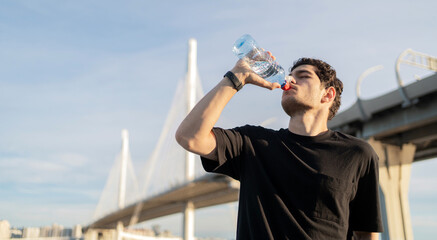 Drinking water from a bottle, a male athlete training in the city in sportswear.