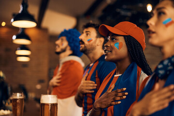 Group of fans with hand on heart singing national anthem while watching world cup in pub. - obrazy, fototapety, plakaty
