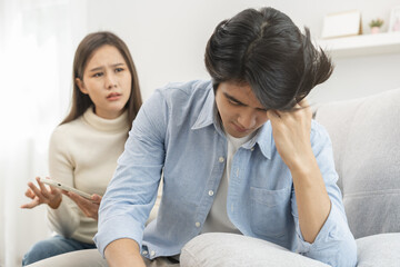 Infidelity, suspicion asian young couple love fight relationship, wife holding cellphone, smartphone cheating on phone, scolding husband about mistrust, distrust and jealousy when sitting at home.