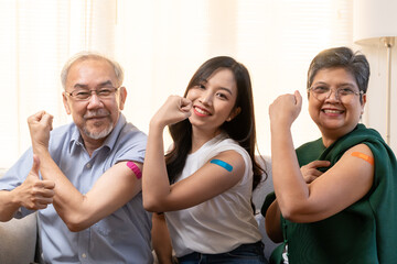 Coronavirus Vaccination, happy asian elderly family with daughter smile strong together, showing bandage on arm protect of covid-19 after injection of vaccine, sitting on couch in living room at home.