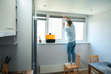 Young man stands on chair and repairs window with screwdriver