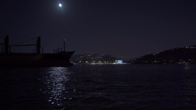 Ship passing through the strait under the moonlight.
Merchant ship sails through the city strait under moonlight at night.
