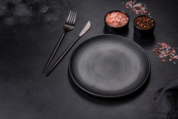 An empty black plate on a dark concrete background with spices, herbs and cutlery