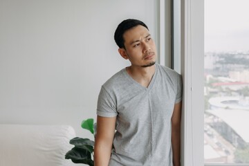 Thinking serious lonely asian man stand lean on the window in his apartment