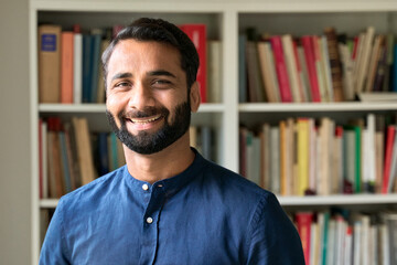 Happy bearded indian professional business man, smiling ethnic teacher, professor or book author standing in front of bookshelves library looking at camera, portrait. Business education concept