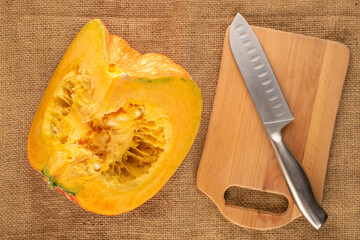 One piece of ripe organic pumpkin with metal knife and separate wooden board on jute cloth, macro, top view.