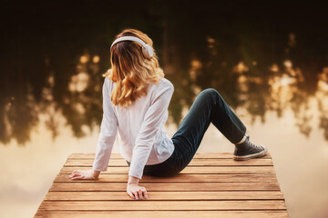 A beautiful blonde woman in a turtleneck and jeans is sitting on a wooden pier with headphones and...