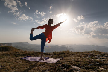Woman doing yoga, Natarajasana asana - outdoor dance pose at sunset in the mountains. A yogi on a rug balances near a cliff during the day