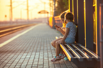 two little girls tourist sisters are sitting on the bench of the railway station and watching the...