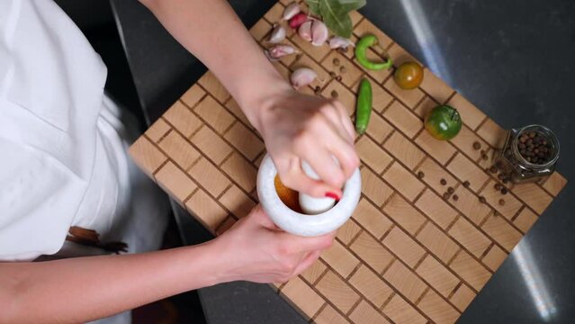 White stone grinding bowl for crushing mixed spices and herbs, granite mortar and pestle for mixing variety of aromatic flavours. Top view shot of woman hands crushing spices for meat and fish