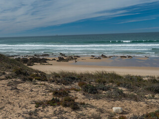 View of empty small sand beach with ocean waves and sharp rocks, stones and green vegetation at wild Rota Vicentina coast near Porto Covo, Portugal.