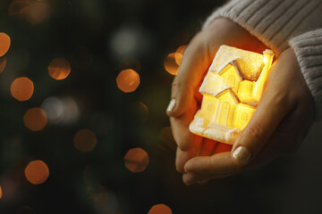 Little glowing house in hands on background of illuminated christmas tree lights bokeh. Magical...