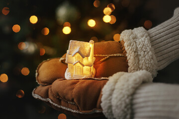 Fototapeta na wymiar Hands in gloves holding little glowing house on background of illuminated christmas tree lights. Magical winter time, atmospheric image. Cozy home. Merry Christmas and Happy Holidays!