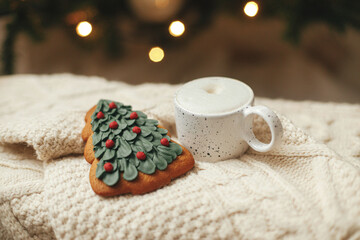 Atmospheric winter hygge. Warm cup of coffee and christmas tree gingerbread cookie on knitted sweater on background of christmas tree with lights. Christmas banner. Cozy holidays