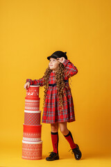 little girl in hat and plaid dress plays with Christmas gift boxes, building pyramid on yellow background in studio. the child is happy to receive many gifts for Christmas. Advertising and discounts.