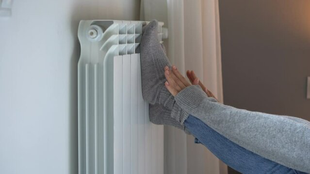 Female person puts feet in grey woolen socks on room central heating radiator. Owner gets cold and tries to warm up feet and hands in uncomfortable apartment in winter. Utility services problems