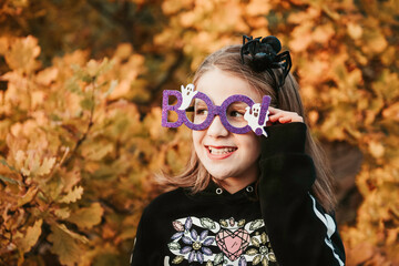 Portrait of cute smiling caucasian teen girl with loose hair wearing black hoodie, violet boo glasses and headband with black spider. She is standing outdoors in autumn park. Halloween decoration.
