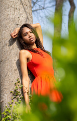 Joven colombiana delgada, mulata con vestido rojo y pelo largo oscuro, apoyada en un tronco con la mano arriba y hojas verdes en la primavera de 2021. - obrazy, fototapety, plakaty