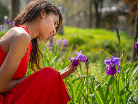 Joven adolescente colombiana de color oscuro con vestido rojo de tirantes, agachada mirando unas flores moradas, en un prado verde exterior , en la primavera de 2021