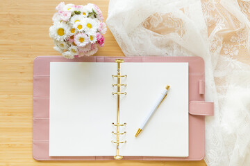 Flat lay, top view of a pastel pink diary, flower bouquet and stationery on a wooden desk.