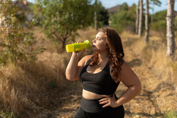 young overweight woman drinking water after jogging in the forest. Portrait of young plus-size...