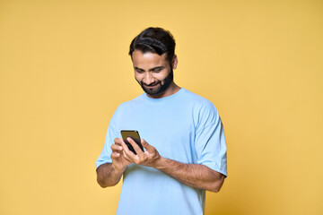 Happy indian man using cell mobile phone isolated on yellow background. Smiling ethnic guy holding smartphone playing game in app, paying online, betting, buying in e commerce shop on cellphone.