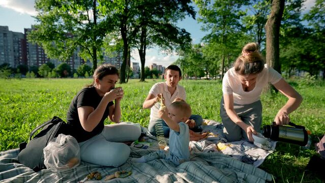 Family members hold picnic in city park. Relatives sit drinking tea on plaid put on grass lawn against tress . Toddler boys enjoy picnic playing on rug
