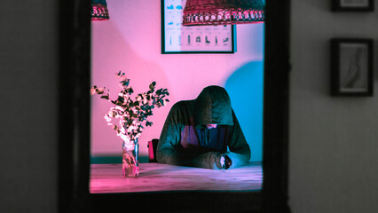 Portrait of a caucasian man sitting on a chair and looking spooky wearing a hoodie. Selective lighting over the man gives a terrorific atmosphere to the scene while he reflects on a mirror. 