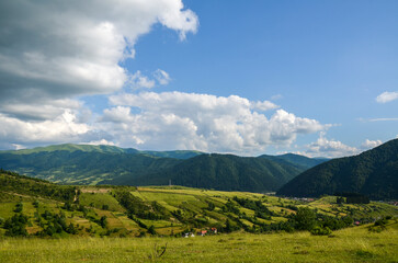 Countryside landscape with grassy pasture on the rolling hills and village in the valley near mountain ridge in sunny summer day. Carpathian Mountains, Ukraine