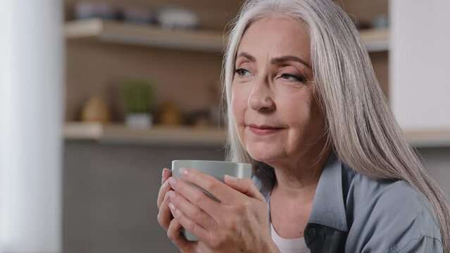 Dreamy mature woman holding cup of hot beverage warming hands on mug preventing flu looking away drinking morning coffee positive dreaming pleasant memories happy elderly grandmother relaxing at home