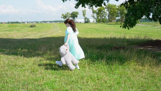 Young pregnant woman in blue dress with a big teddy bear walking in the park.