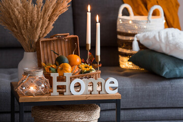 Cozy autumn concept. Home warmth in cold weather. Still-life. Pumpkins, wicker basket, candles, flowers and the inscription home on the coffee table in the home interior of the living room.