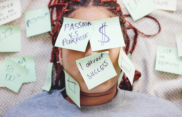 Black woman tired with sticky note on face, motivation for success and money on bed. African girl in bedroom, stick idea in writing on skin and eyes, for manifestation of dreams of house and wealth