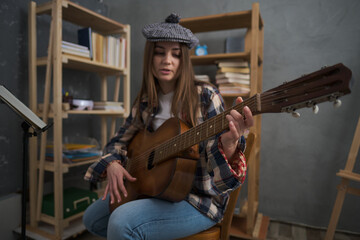 Obraz na płótnie Canvas A cute girl in a fashionable beret plays a classical guitar while sitting in a room. Many books are behind on the shelves. Theme of study, student, music, hobby and spending free time at home
