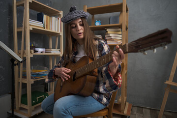 A cute girl in a fashionable beret plays a classical guitar while sitting in a room. Many books are behind on the shelves. Theme of study, student, music, hobby and spending free time at home
