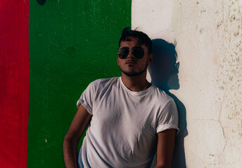 Hombre joven modelo con gafas de sol, gorra y camisa blanca apoyado en un muro de colores 
