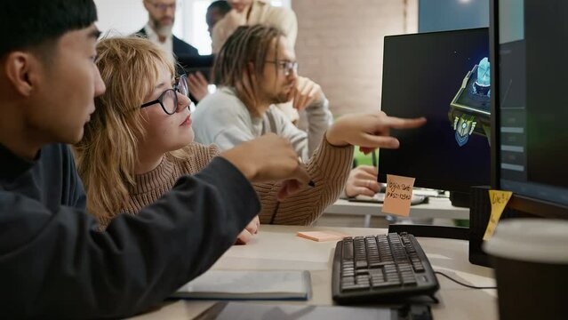 A European woman and an Asian male artist discuss the 3D model in a computer game and application development studio among other team members.
