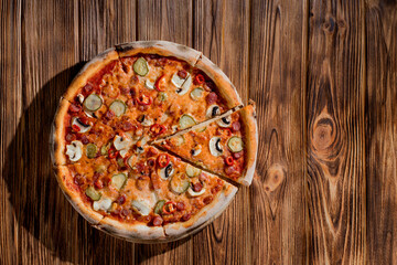 Appetizing pizza with hunting sausages, mozzarella cheese, pickled cucumbers, chili peppers on a tomato base on wood background