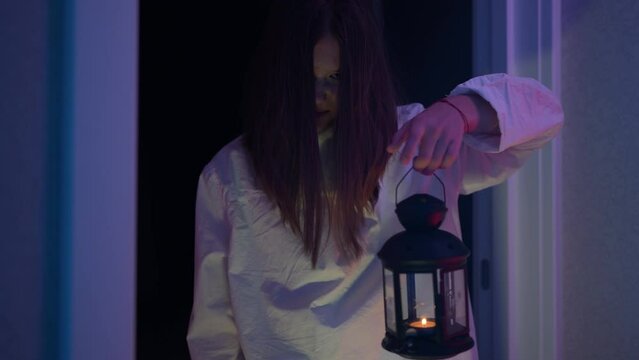 A little girl in a ghostly white halloween sundress holds a lantern with a candle, scaring someone in the hallway of a haunted house. 4k footage