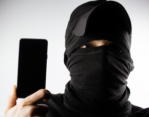 A masked criminal with a smartphone in his hand. A criminal in a balaclava holds a smartphone for copy space. Theft of a smartphone, personal data.