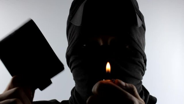 A criminal, a thief in a mask, a balaclava with a wallet in his hand and a match. The concept of stealing money, wallet. A pickpocket thief in black.