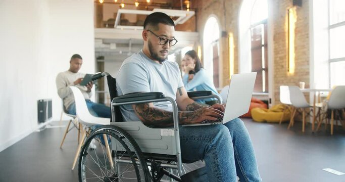 Latin man in wheelchair works on laptop against colleagues