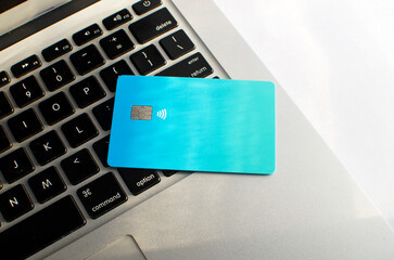 Credit card with visible chip and laptop, on top of a table with soft lights and shadows. Blue color card on white surface. Concept: finance, purchases, payments, loan, spending, investments.