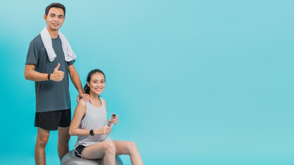 Asian beautiful happy woman holding water bottle and sitting on fit ball with boyfriend after exercise isolated on blue color background.Concept of slim and healthy girl workout.