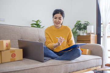 Business Asian women sit on sofa use laptop computer checking customer order online shipping box at home. Starting Small business entrepreneur SME freelance. Online business, Work at home SME concept.