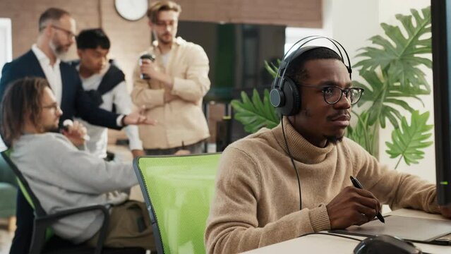 African American male artist designer in headphones draws on tablet 3D models for games. Against background, multinational team of developers are discussing concept of presentation of project.